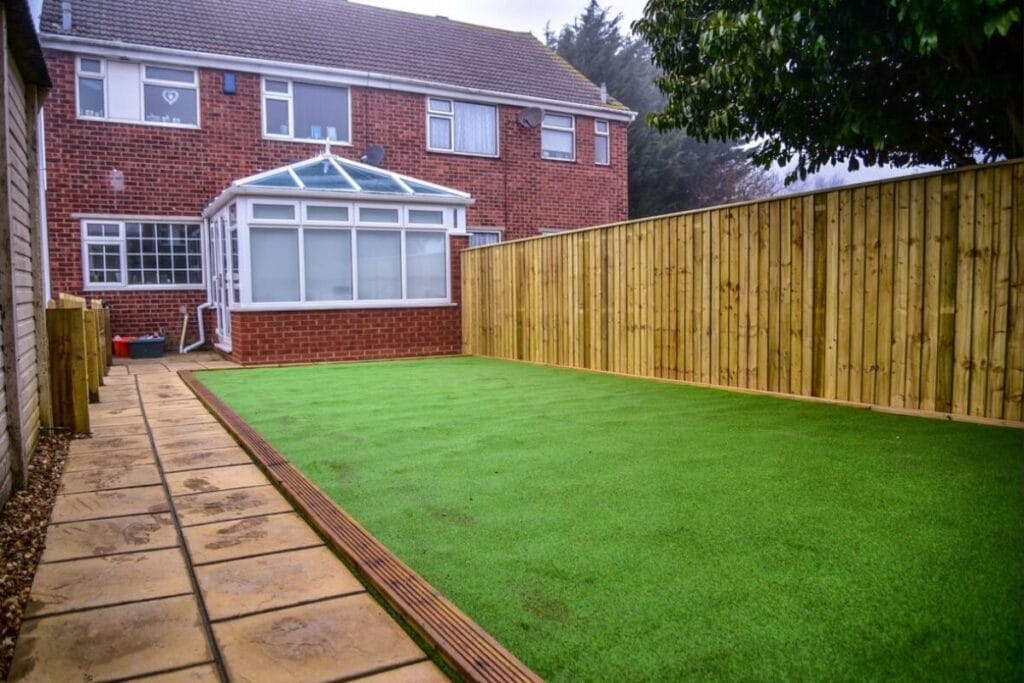 Wooden fence enclosing a back garden with artificial grass and a red-brick house featuring a conservatory.