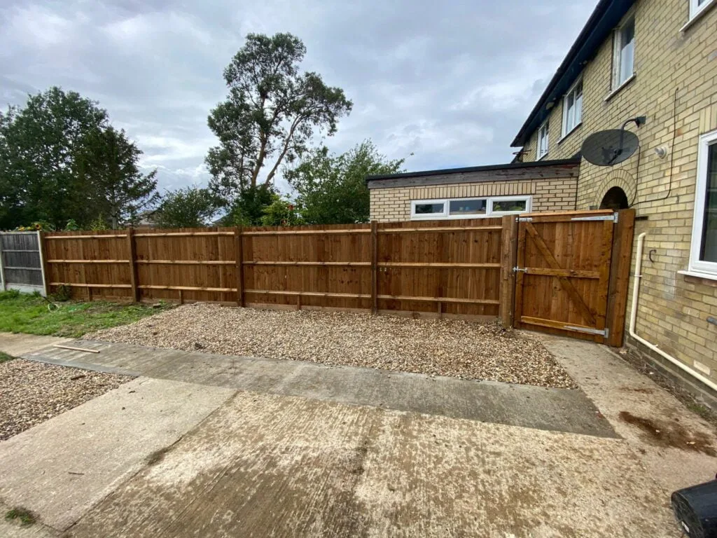 Wooden fence with horizontal rails and a matching wooden gate enclosing a gravel driveway beside a brick house.